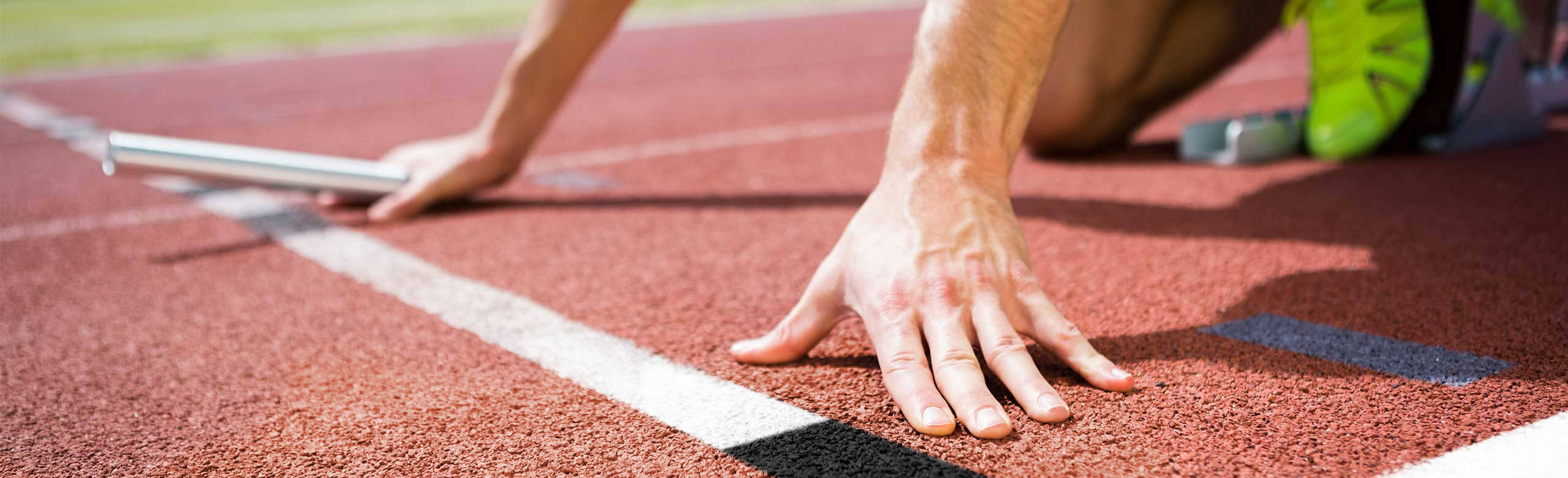 Close-up of an athlete in starting position on a red synthetic track with relay baton in hand and spikes in the block.
