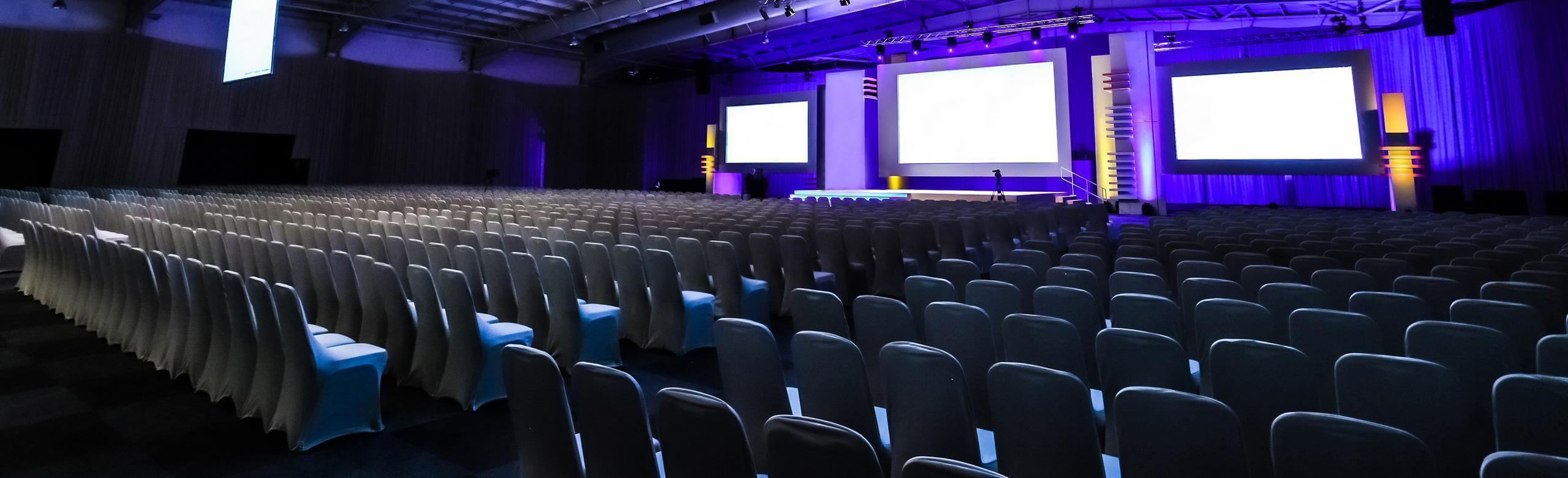 Wiele rzędów krzeseł skierowanych w stronę sceny. Wiele rzędów krzeseł skierowanych w stronę sceny. Scena znajduje się w hali sportowej i jest podświetlona fioletowym światłem. Trzy duże ekrany wiszą na ścianie sceny.