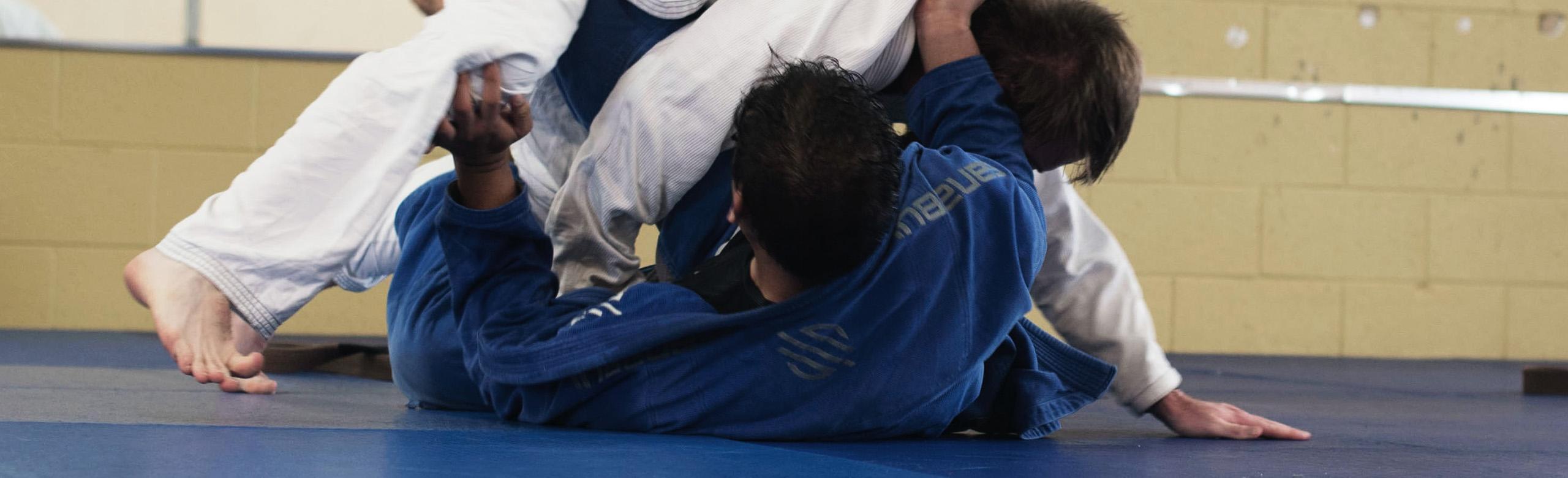 Dwie osoby ubrane w stroje do jiu-jitsu walczą w parterze na niebieskich matach w hali sportowej.