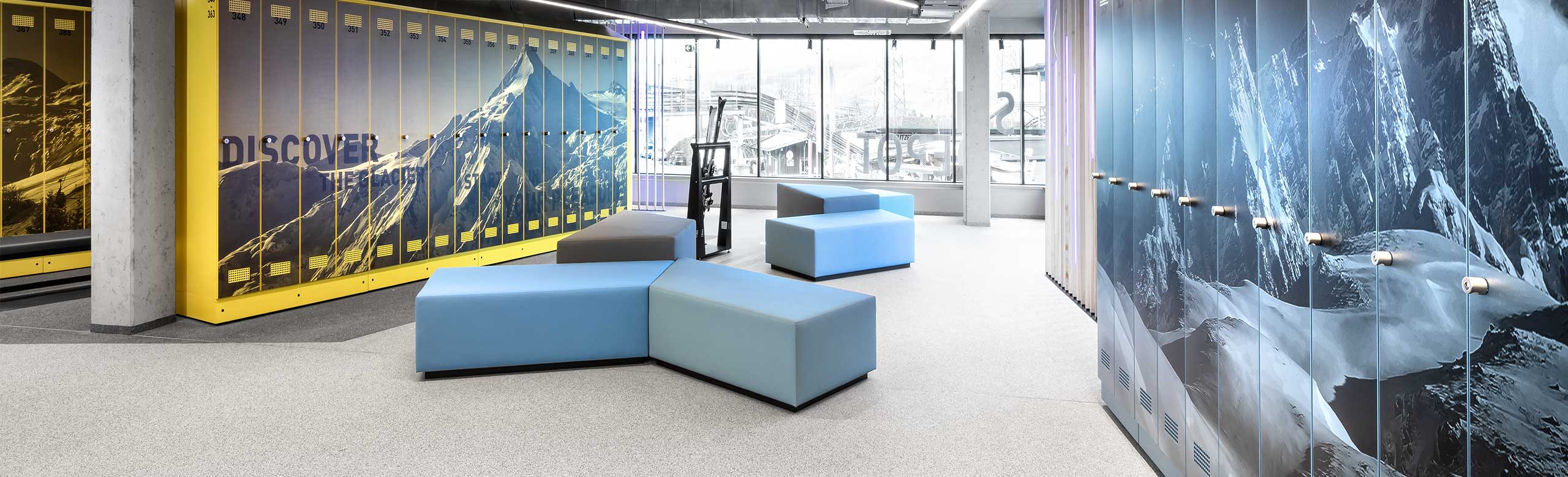 Jasno oświetlona przechowalnia sprzętu narciarskiego na górskiej stacji kolejowej. Jasno oświetlona przechowalnia sprzętu narciarskiego na górskiej stacji kolejowej. Na końcu pomieszczenia znajduje się duże okno frontowe, po prawej i lewej stronie obrazu widać szafki z motywami górskimi. Pośrodku znajduje się kilka foteli w kolorze niebieskim i szarym. Podłoga jest jasna, a poniżej szafek ciemnoszara.